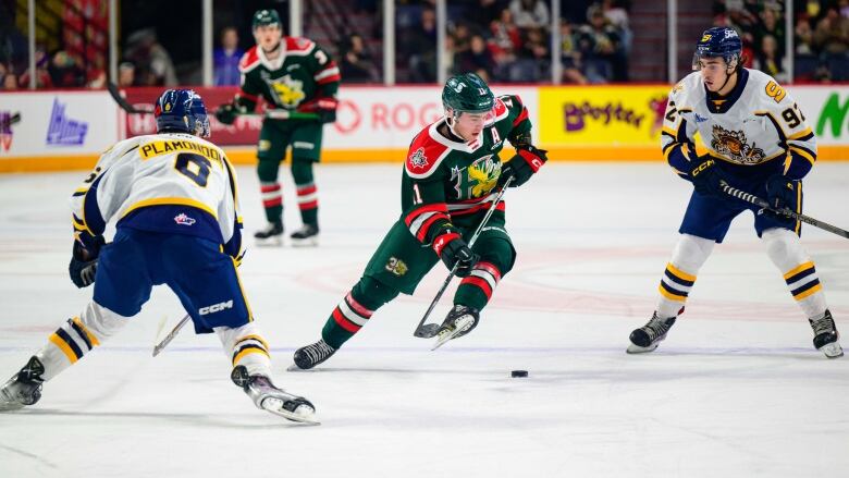 A junior hockey player wearing a green and red jersey tries to squeeze between two opposing players.