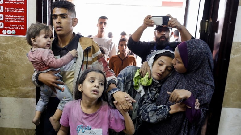 A group of five people walk into a hospital, including, a young man holding a baby, a girl, and a woman who is supported by another woman.