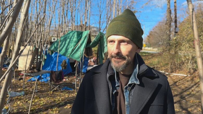 A man stands in front of a shelter made of tarps in a wooded area. He is wearing a toque and several layers under his coat.