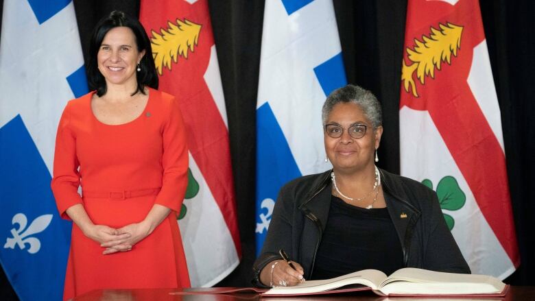 Two women, one standing, one seated with a pen in her hand, in front of flags.