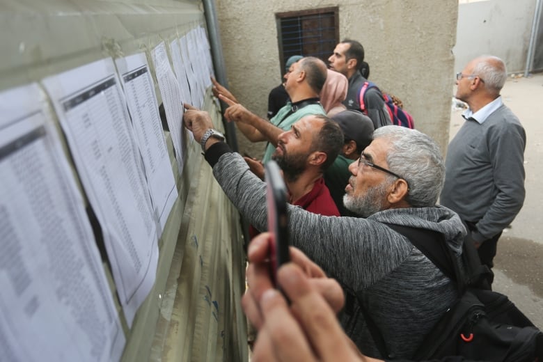 A group of people read a list of names taped to a wall. 