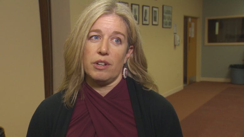 A women with blond hair and blue eyes. She is wearing a dark red shirt, a black cardigan and beaded earrings.