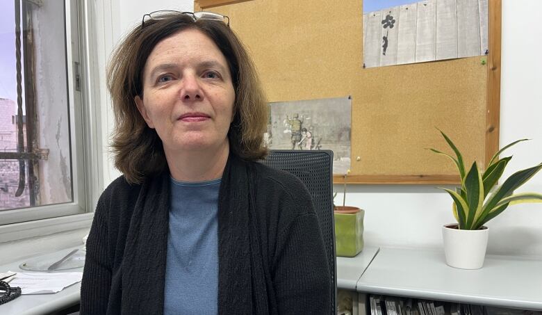 A woman with shoulder-length brown hair sits on a chair in an office, a potted plant to the right in the photo. She has glasses on the top of her forehead and wears a black sweater over a blue shirt.