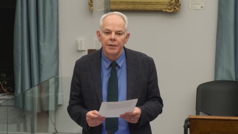 A man in a dark suit and checkered green tie standing while posing questions to the premier.