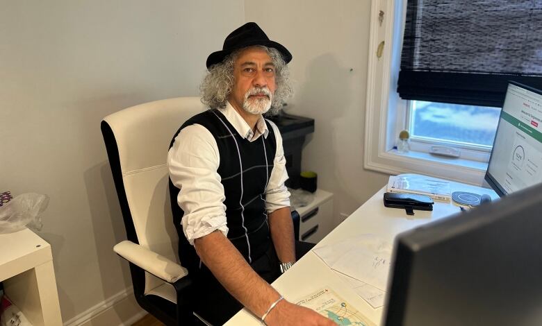 A man in a hat and vest sits at a desk with a computer, posing for a photo.