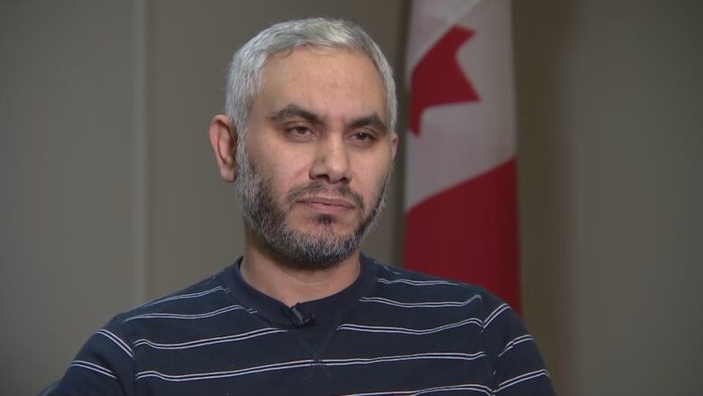 A bearded man sits in front of Canadian flag.