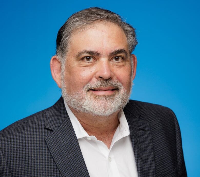 A man with grey hair and a white beard poses for a professional headshot while wearing a suit jacket and white shirt.