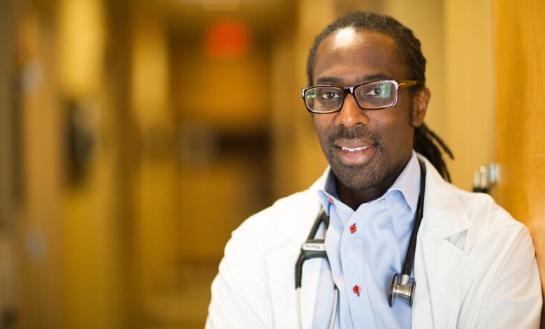 A man wearing a white coat and stethoscope is seen standing in a hallway.