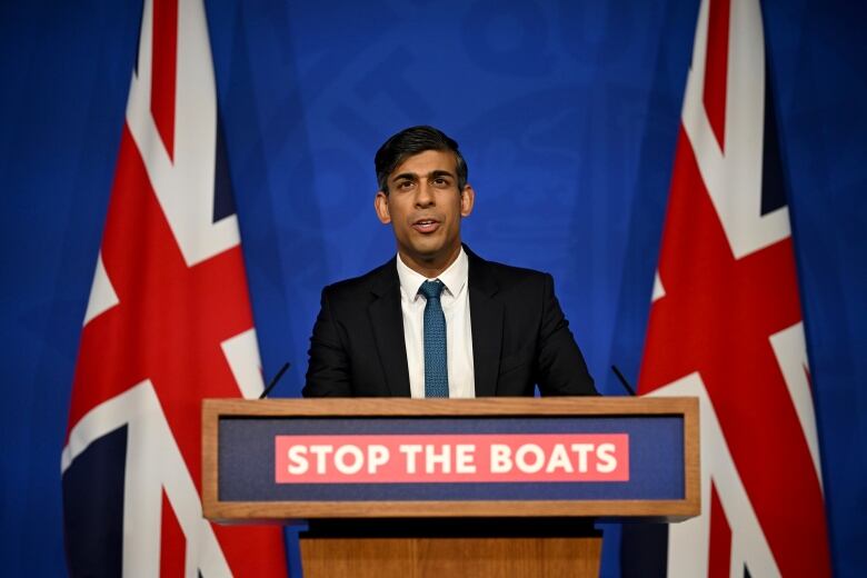 A man wearing a black suit and a blue tie stands at a podium that says 