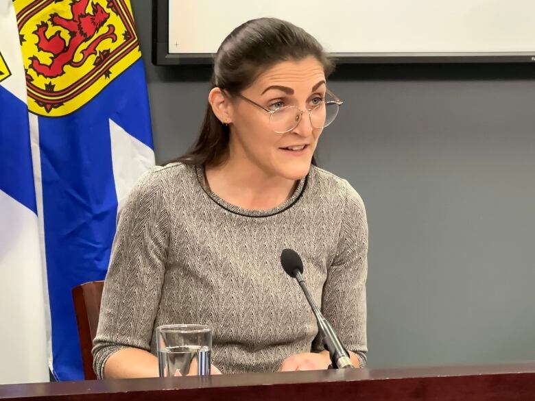 A woman in a grey sweater and glasses speaks into a microphone.