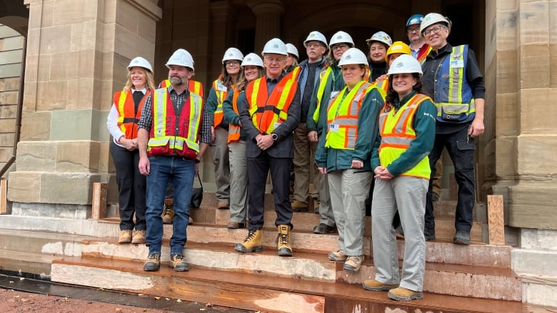 Fifteen smiling people with safety vests and hard hats stand on the steps of a stone building.