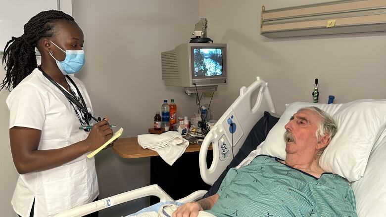 A nurse is shown wearing a stethoscope around her neck and writing on a yellow notepad as she speaks to a hospital patient who is in bed.