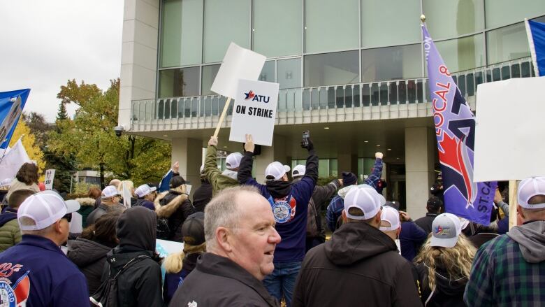 A crowd (seen from behind) holds picket signs in front of a tall city building.