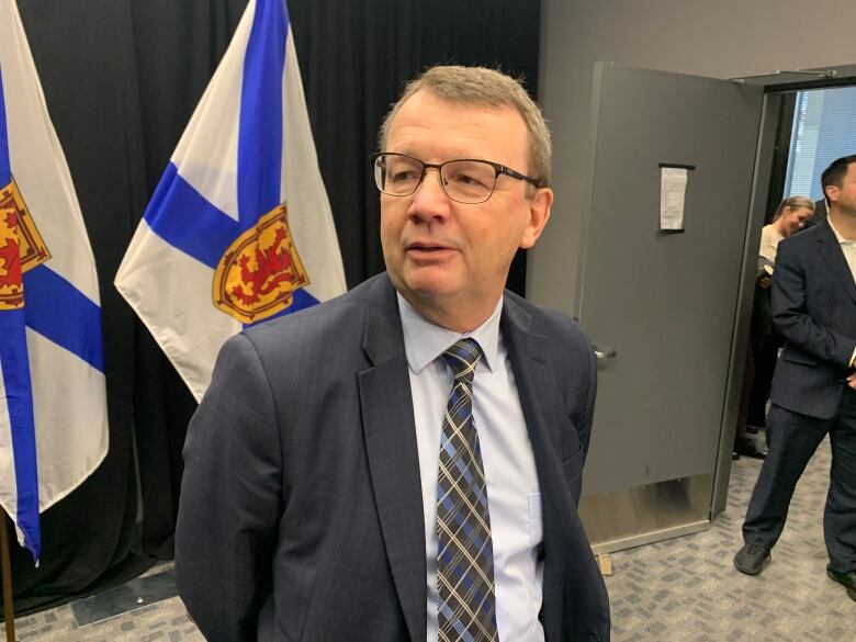 A man wearing a suit stands in front of a Nova Scotia flag.