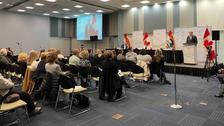 Rows of people sit on chairs facing the front of the room. Dr Michael Gardam is standing at a podium speaking into the microphone. 