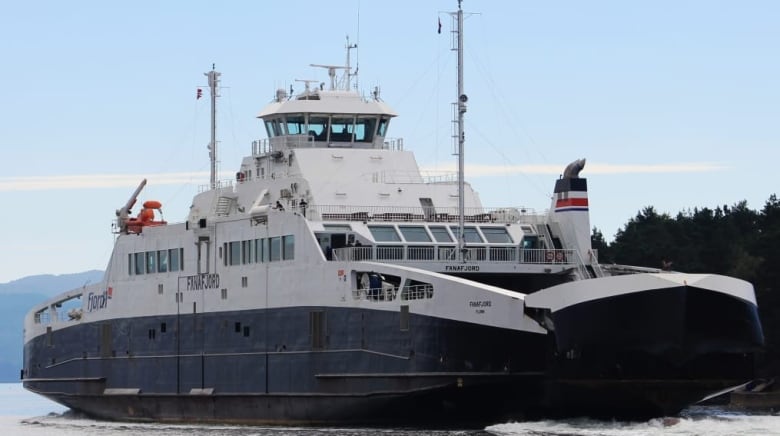 A large ferry with its nose cone opening to let vehicles drive off is shown approaching a dock. 