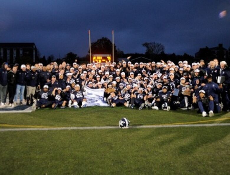 A team of football players sit, kneel and stand together beneath the uprights at night. Players in the middle hold a championship banner.