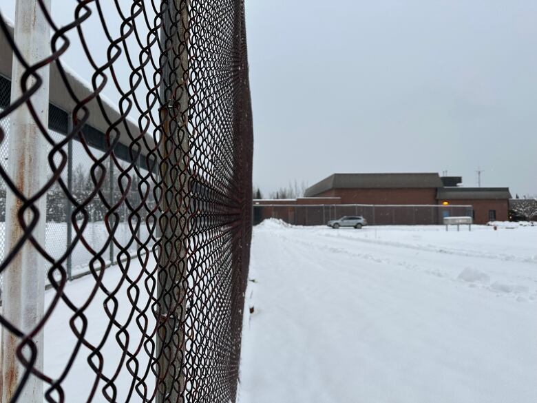 A large brick building is seen in the distance behind a chain link fence. There is snow on the ground.