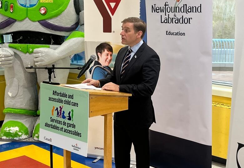 A man stands at a podium in front of three banners. A large statue of Buzz Lightyear as tall as the room itself stands in the background.