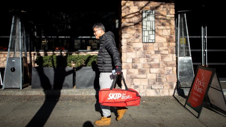 A delivery worker carries a red bag reading SkiptheDishes past a restaurant.