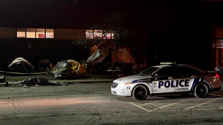 A police car at night in front of tents, taped off by police tape.