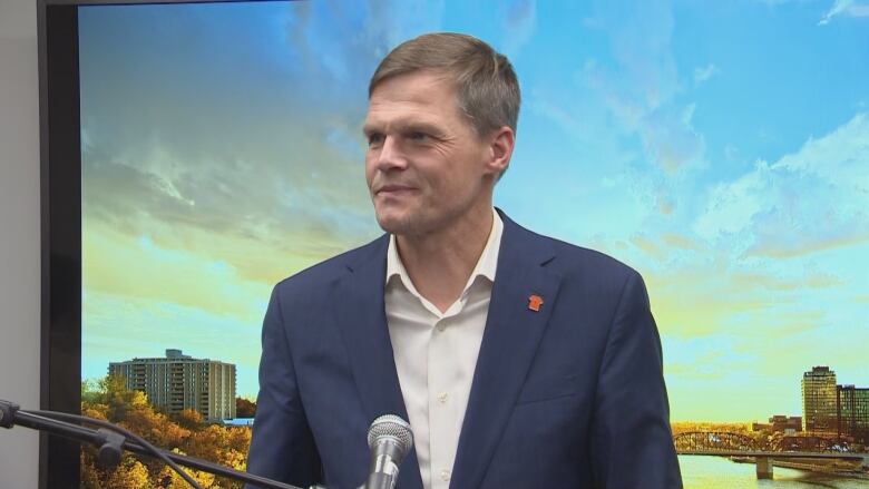 A white man with short brown hair is wearing a blue dress coat over a white dress shirt. He is standing in front of a photo of a sunset.