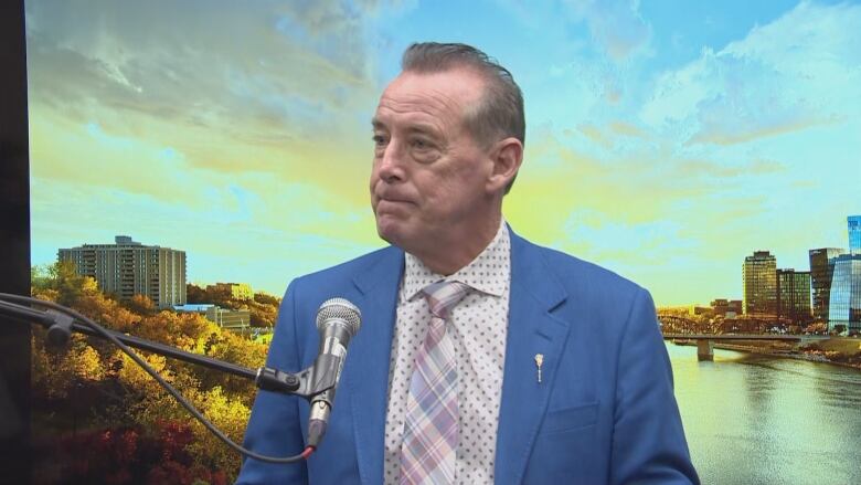 An older white man with thinning grey hair is wearing a blue dress coat over a white patterened shirt. He is wearing a plaid tie. The man is standing at a microphone; behind him is a photo of a sunsetting over a city.