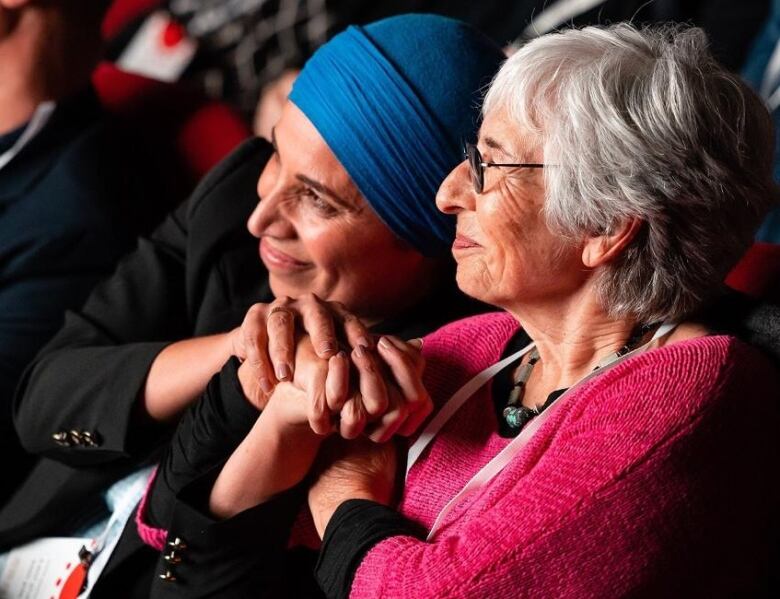 Two women sit together in a crowd, smiling brightly, holding hands, and leaning into each other. 