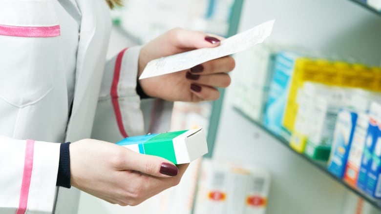 A closeup shows the hands of a woman in a white lab coat in a store, holding a prescription in one hand and a box of medicine in her other. 