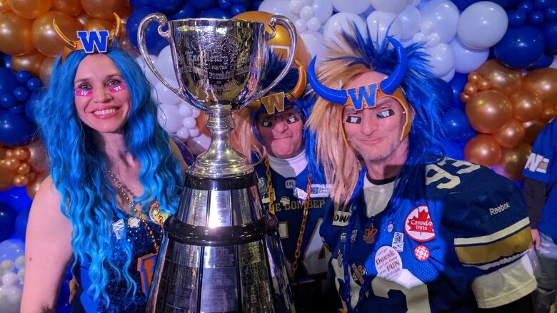 Three people dressed up in Winnipeg Blue Bomber regalia and a silver trophy.