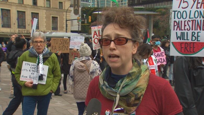 A woman speaks to a camera in the background of a protest.