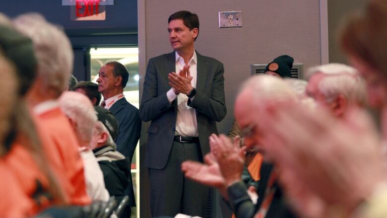 A tall white man claps while looking sideways, while in the background of dozens of people clapping.