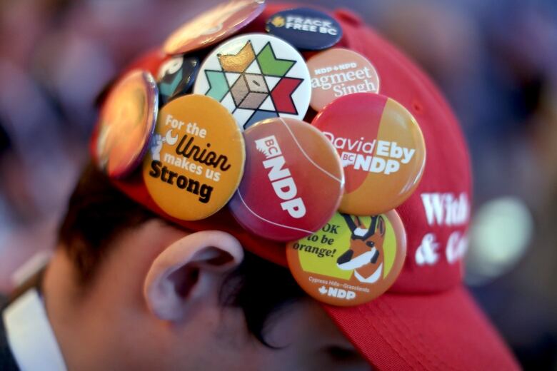 A man wears numerous pins on his hat, most of them orange and supporting the New Democratic Party.