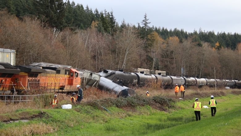 A train is tipped over onto the grass beside the track and several people wearing high visibility vests stand around. 