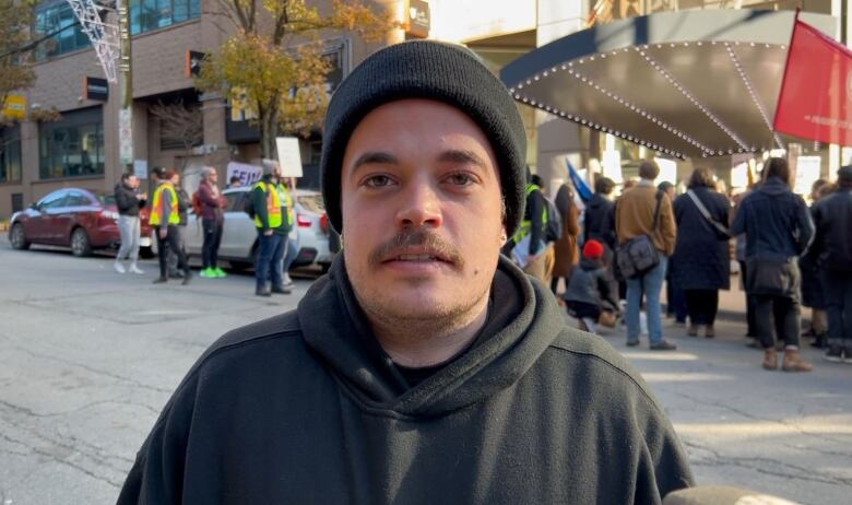 A man in a black hoodie and a black toque speaks to the camera