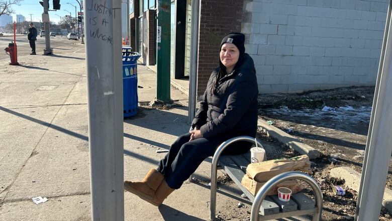 A woman is sitting on a bench inside a bus shelter that is missing all of its glass.