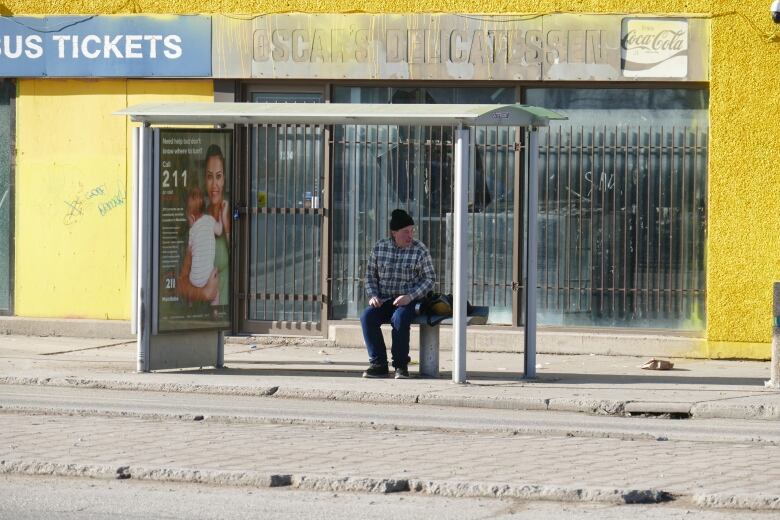 A man is sitting on a bench in a bus shelter that is missing all of its panes of glass.