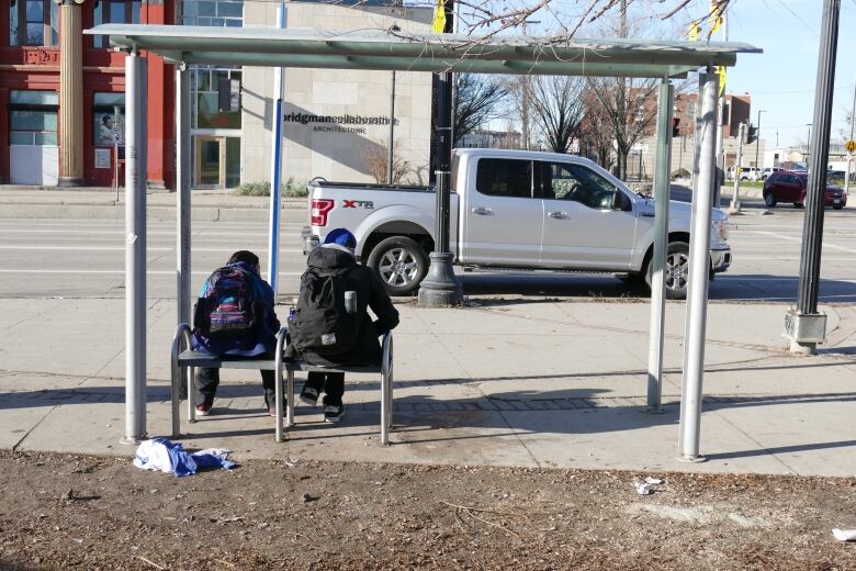 Two people are sitting in a bus shelter that is missing all of its glass.