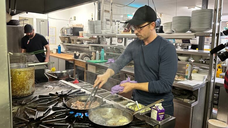 A man wearing a ball cap is in a restaurant kitchen at the stove working two frying pans. 