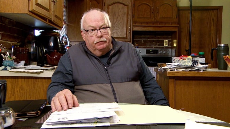 A man sits at his kitchen table. He's a larger man with a white mustache. There is a folder of papers in front of him. Behind him you can see oak kitchen cabinets.