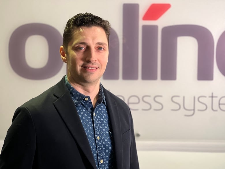 A man stands in front of a sign that says Online Business Systems. He's wearing a navy jacket and a blue button-up shirt.