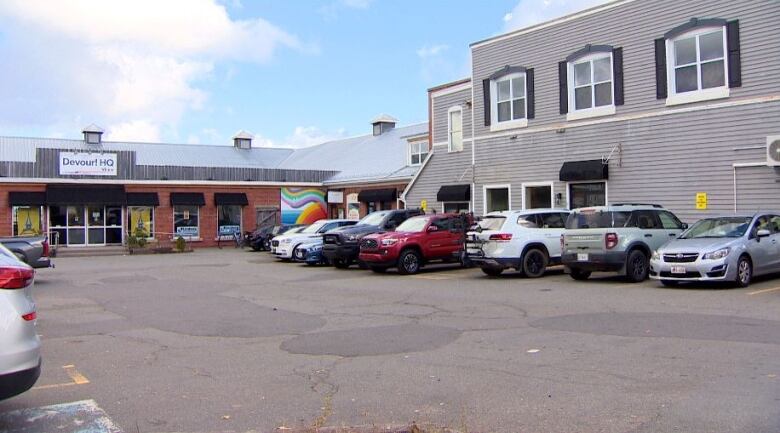 A parking lot in the foreground contains cars while a one-story brick building has a sign for Devour! on it. The Devour building is the long part of the L shaped building, with the foot the apartment building on the right