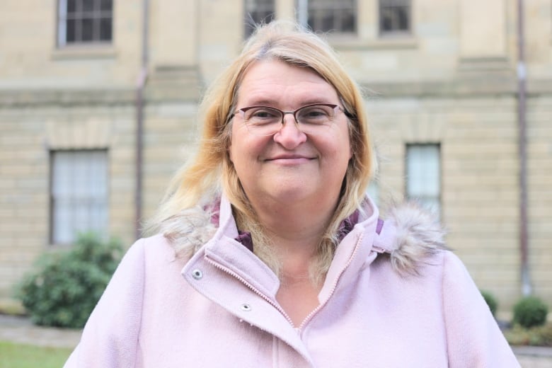 A woman with glasses stands in front of Province House in Nova Scotia. She is wearing a jacket with a hood.