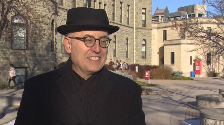 A man stands smiling on University of Winnipeg campus, wearing round, black glasses and a black felt, brimmed hat