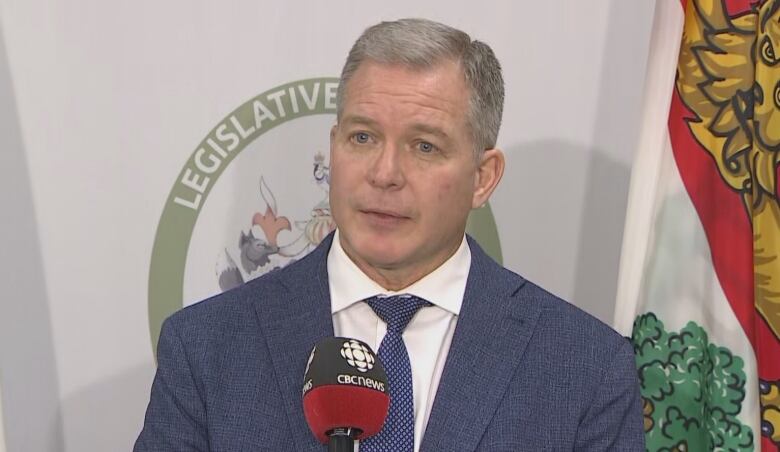 A man with short grey hair in a dark blue suit stands in front of a microphone.