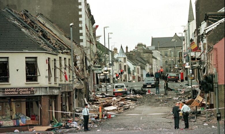 People stand on a street strewn with boards and debris from buildings damaged by a bomb.