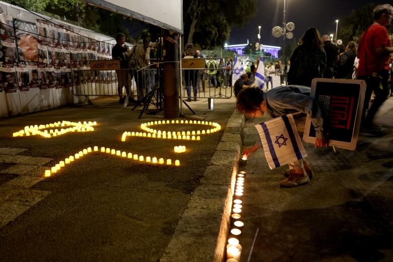 A woman holding a blue and white Israeli flag bends to place a small candle on the ground in front of other candles arranged in the date 7-10 and in the shape of a Star of David.