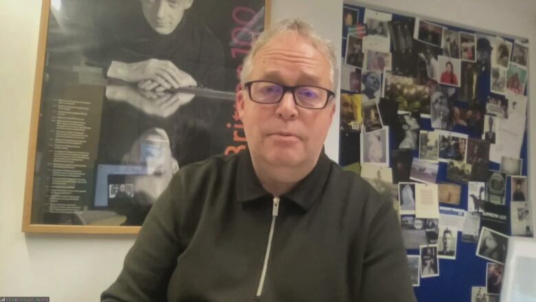 A man wearing a zip-up black sweater and black-rimmed glasses. He sits in front of a poster and a bulletin of photos.