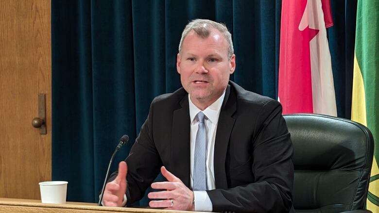 A white man with thinning grey hair is wearing a black suit jacket over a white dress shirt with a light blue tie. He is sitting in a leather desk chair, in front of some dark curtains and flags. 