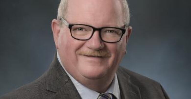 Man picture from chest up, wearing a grey suit facing camera in professional headshot. Has grey hair, moustache and glasses. 